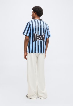 Homme aux cheveux bouclés portant une chemise à manches courtes à rayures verticales bleues et noires avec le texte « HEATH'S TEAM » au dos et un pantalon blanc à jambes larges.