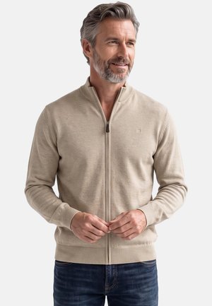 Middle-aged man with gray hair and beard wearing a beige zip-up sweater and dark jeans, looking slightly to the side and smiling.