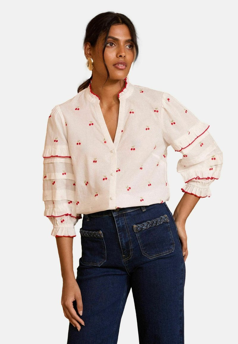 Blusa bianca con ricami di ciliegie rosse e maniche arricciate, abbinata a jeans in denim scuro con dettagli delle tasche intrecciati.