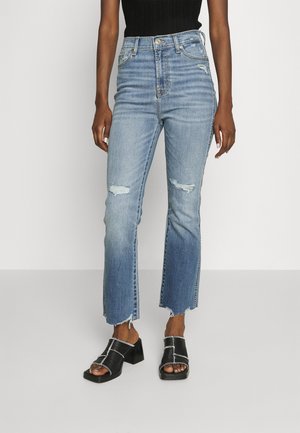 Jeans high-waisted di un blu chiaro con toppe strappate e orli grezzi, abbinati a sandali neri con tacco a blocco e dettagli di cucitura bianca.