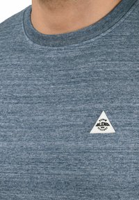 Sweatshirt albastru heather cu guler rotund, guler ribat și un etichetă albă triangulară cu text negru pe zona stângă a pieptului.