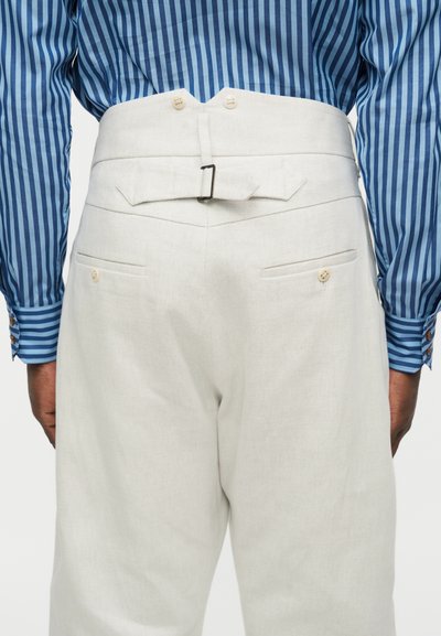 Άνδρας που φοράει λευκό παντελόνι με ψηλή μέση, ρυθμιζόμενη πίσω τιράντα και τσέπες με κουμπιά, συνδυασμένο με μπλε ριγέ μακρυμάνικο πουκάμισο.