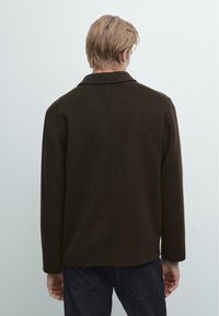 Giacca marrone con una vestibilità rilassata, colletto a punta e maniche lunghe. Texture liscia, design sul retro senza adornamenti, dettagli di cucitura minimal.