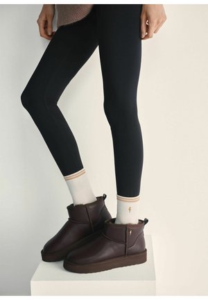 Brązowe skórzane botki za kostkę z futrzanym wykończeniem, zestawione z czarnymi legginsami i białymi skarpetkami z złotymi akcentami. Ułożone na jasnym tle.