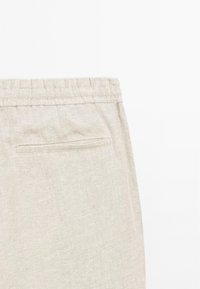 Pantalon en lin beige avec une taille élastique et une poche passepoilée unique à l'arrière côté droit, présenté sur fond blanc.