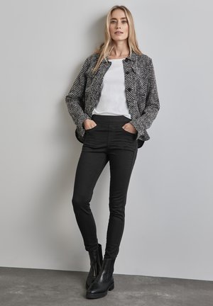 Veste noire texturée sur une chemise blanche, associée à un pantalon noir ajusté et des bottines. Les caractéristiques incluent des boutons et des poches.
