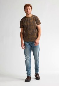 Bruine camo T-shirt met korte mouwen en een ronde halslijn, gecombineerd met lichtblauwe jeans met slijtage-effect en donkerbruine veterlaarzen.