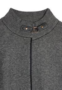 Grå tekstil med et strukturert mønster, med en svart lærdetalj og et klips i gullfarge ved kragefestet.