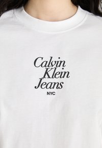 T-shirt alb din bumbac, cu text negru care spune "Calvin Klein Jeans NYC" într-un font îndrăzneț și modern. Stil cu gât rotund.