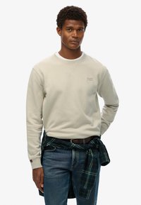 Sudadera gris claro con cuello redondo y puños acanalados, combinada con jeans azules. Una camisa de cuadros verde y negra está atada a la cintura.