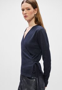 Granatowy sweter zapinany na bok z długimi rękawami, o teksturowanym wykończeniu i bocznym wiązaniu. Materiał ma subtelny połysk.