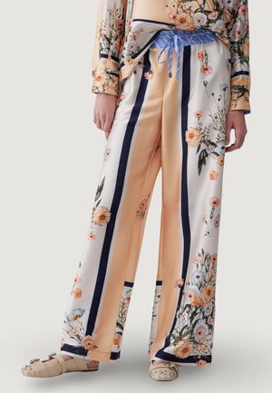 Weit geschnittene Satinhosen mit vertikalen marineblauen Streifen, pfirsichfarbenem Hintergrund und Blumenmuster, kombiniert mit verzierten beigen Sandalen.