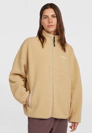 Vrouw met bruin haar die een beige fleecejack met rits en een bruine broek draagt, staand met één hand in de jaszak tegen een effen achtergrond.