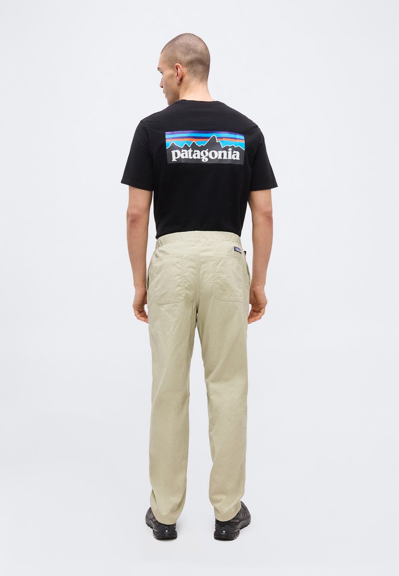 Person facing away wearing black Patagonia t-shirt with logo and beige pants, standing against a plain light background.