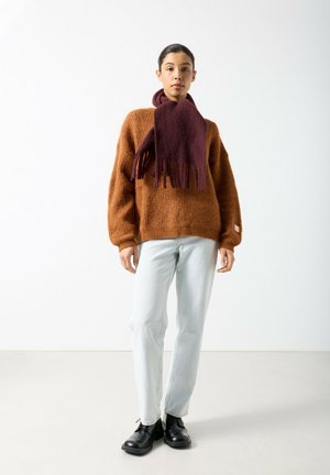 Brown knitted sweater, wide sleeves, paired with light blue jeans. A burgundy scarf with fringe detail complements black shoes.