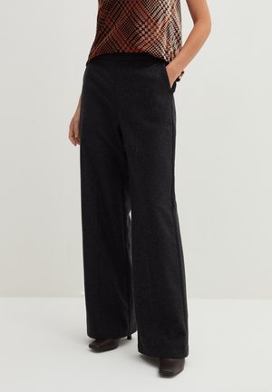 Woman wearing dark gray wide-leg pants with hands in pockets, paired with brown heeled shoes and a patterned top.