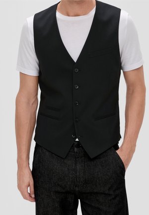 Homme portant un gilet noir boutonné sur un t-shirt blanc à manches courtes et un jean sombre, debout avec la main droite dans la poche devant un fond uni.