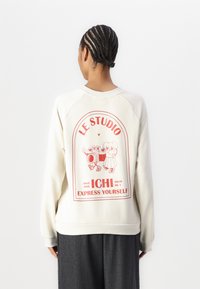 Sudadera de color crema con un gráfico en la parte posterior que presenta texto rojo y personajes de dibujos animados. Textura suave, corte relajado y cuello y puños de canalé.
