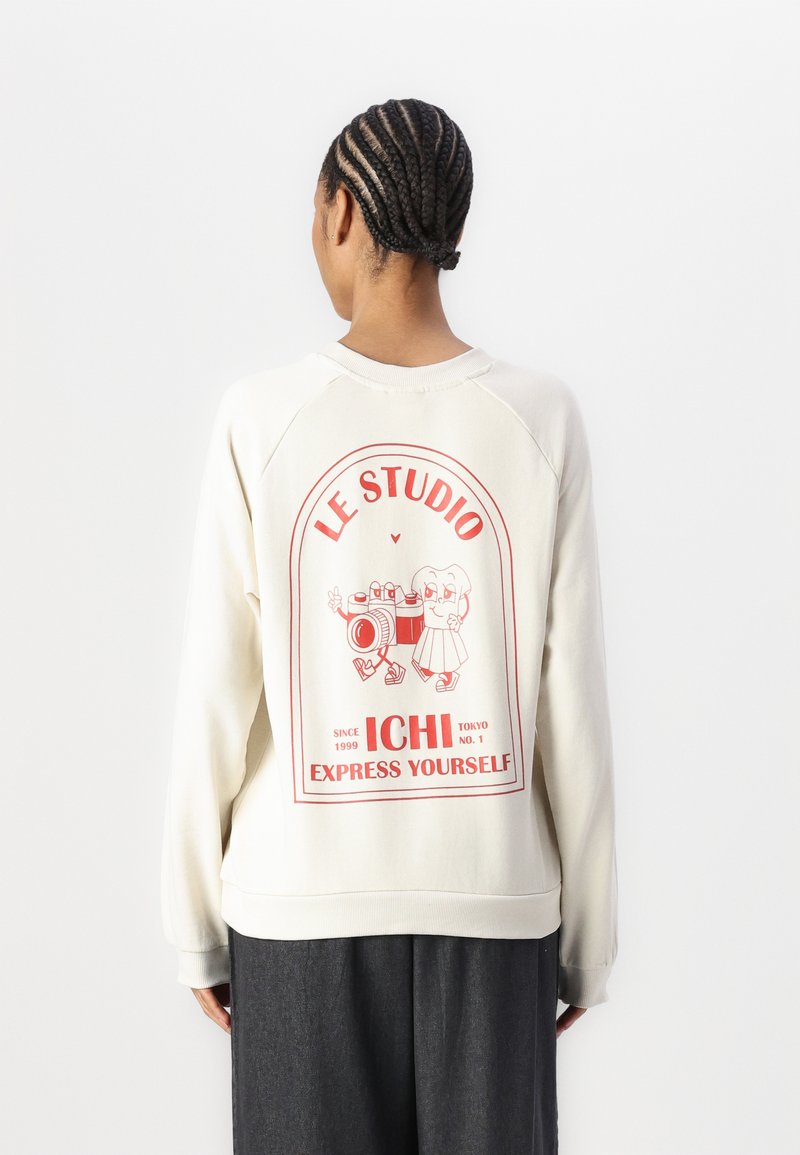 Sudadera de color crema con un gráfico en la parte posterior que presenta texto rojo y personajes de dibujos animados. Textura suave, corte relajado y cuello y puños de canalé.