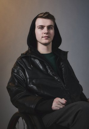 Schwarze Lederjacke mit Reißverschluss und Kapuze, getragen über einem grünen Hemd. Die Person sitzt im Rollstuhl, vor einem neutralen Hintergrund mit sanfter Beleuchtung.