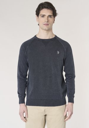 Giovane uomo che indossa un maglione blu scuro a maniche lunghe e pantaloni beige, in piedi e rivolto verso l'obiettivo su uno sfondo semplice.