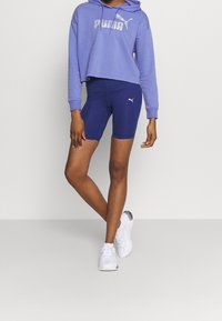 Felpa corta viola con logo bianco "PUMA", abbinata a pantaloni bike blu navy con piccolo logo e scarpe da ginnastica bianche con accenti argento.