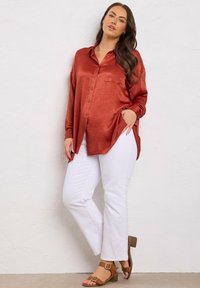 Woman with long dark hair wearing a loose rust-colored satin shirt, white pants, and tan heeled sandals, standing against a white wall.