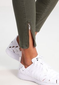 Olivgröna tajta byxor med sidodragkedjor, kombinerat med vita vävda sneakers med utskärning och texturerad ovandel.