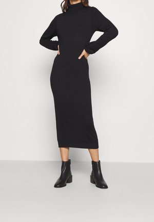 Kobieta stojąca z rękami na biodrach, ubrana w długą czarną sukienkę z golfem i czarne botki na płaskim obcasie na jednolitym tle.