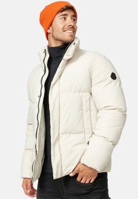 Pufferjakke i off-white med vandret syninger, høj krave, lynlås lukning og en sidelomme. Kombineret med en orange strikket hue.