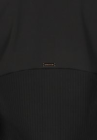 Schwarzes Sportoberteil mit einem glatten oberen Abschnitt und einem strukturierten, gerippten unteren Teil, versehen mit einem kleinen metallischen Logo-Akzent in der Mitte.