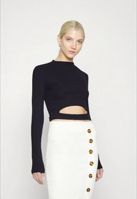 Mulher loira a usar uma blusa preta de manga comprida e curta na altura da cintura, com um corte na frente, e uma saia branca de cintura alta com botões de tartaruga.