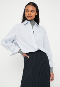 Koszula na guziki z długim rękawem w jasnoniebieskim kolorze z czerwonymi paskami, zestawiona z długą czarną plisowaną spódnicą. posiada kieszeń na piersi i szerokie mankiety.