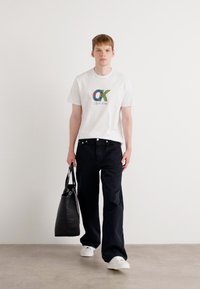 White cotton t-shirt with colorful “CK” logo, paired with black wide-leg jeans. Model holds a black tote bag, wearing white sneakers.