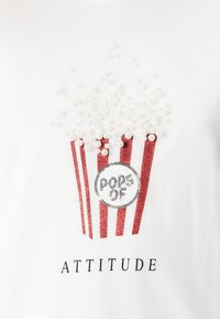 Bílý tričko s grafikou popcornu zobrazující červený pruhovaný kbelík, třpytivé detaily a perlové akcenty pro popcorn. Obsahuje text "ATTITUDE".