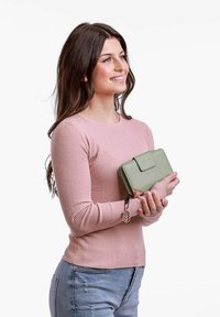 Green leather wallet with a clasp, featuring a patterned interior and a decorative keychain. Model wearing a pink ribbed sweater and blue jeans.