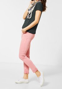 Růžové úzké džíny v kombinaci s tmavě zeleným tričkem a vzorovaným šátkem. Bílé tenisky dokončují outfit. Hladká textura, ležérní styl.