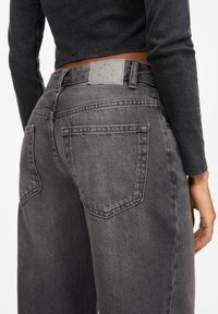 Jean gris taille haute avec un effet délavé, grandes poches arrière et un patch en cuir embossé du logo sur la ceinture.