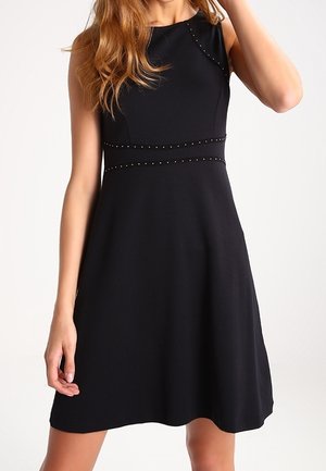 Ærmeløs sort kjole med subtile nitte-detaljer ved taljen og øverste del af brystet, båret af en kvinde med bølget hår, lys baggrund.