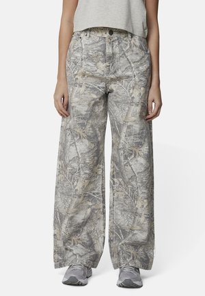 Osoba nosząca spodnie z wysokim stanem i szerokimi nogawkami z szarym i beżowym wzorem gałęzi drzewa, w połączeniu z szarymi trampkami i krótkim, jasnoszarym topem.