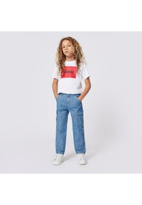 Vit t-shirt med röd rektangel och svart "HUGO"-text, kombinerad med blå cargobyxor. Vita sneakers fullbordar outfiten.