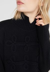Pull noir en maille avec un col montant et des boucles texturées dans le motif. Il comporte des manches longues et un motif côtelé sur l'ensemble.
