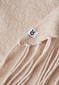 Sciarpa di lana color beige chiaro con una finitura morbida e testurizzata. Presenta frange delicate ai bordi e un'etichetta di marca per un tocco di dettaglio.