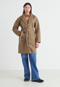 Abrigo marrón texturizado con cuello, cinturón y bolsillos, combinado con jeans acampanados azules y calzado negro. Diseño minimalista con colores neutros.