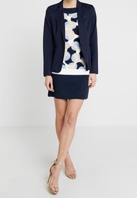 Blazer navy con un patrón texturizado, combinado con una blusa estampada en tonos crema y azul, y una falda navy. Tacones de tiras beige.