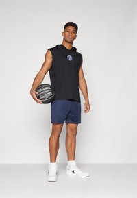 Schwarzes ärmelloses Hoodie, marineblaue Shorts, weiße Sneakers. Hält einen schwarzen Spalding-Basketball. Glatter Stoff, lockere Passform, einfaches Design.