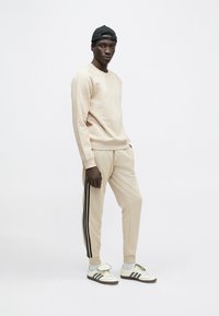 Béžová mikina a zodpovedajúce tepláky s čiernymi bočnými pruhmi. Model nosí čiernu čiapku a biele tenisky s čiernymi pruhmi. Jednoduchý dizajn.