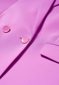 Gros plan sur un blazer violet vif avec deux boutons décoratifs lavande bordés d'or et un rabat de poche sur un tissu lisse.