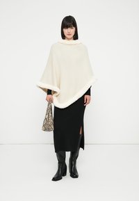 Donna che indossa un poncho bianco con bordi in pelliccia, una lunga gonna nera con spacco, stivali neri, mentre tiene una borsa con motivo in pelle di serpente.