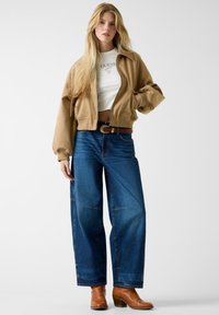 Bijgesneden bruine bomberjack, wit T-shirt met logo, brede blauwe jeans, bruine riem en bruine enkellaarsjes. Soepele textuur en casual pasvorm.